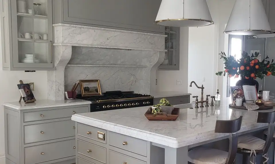 White veined stone island and carved stone range hood surround over a black and brass range with sage gray cabinets, a kitchen from Melling Granite, a source for granite countertops in Cedar City