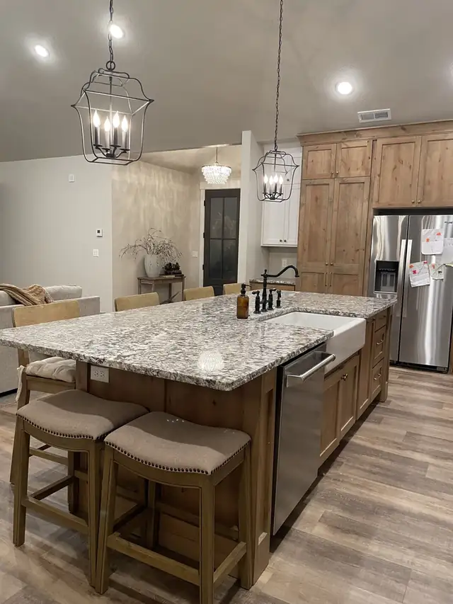 Kitchen island with a speckled granite top, farmhouse sink and wood bar stools under lantern pendants
